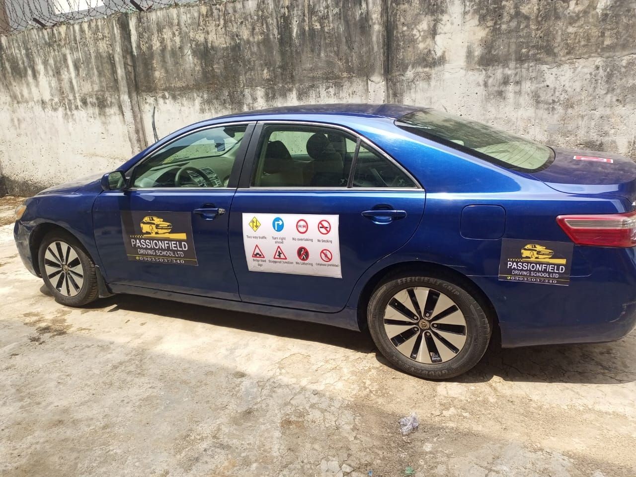 Blue car marked with Passionfield Driving School logo and safety signs, parked against a concrete wall.